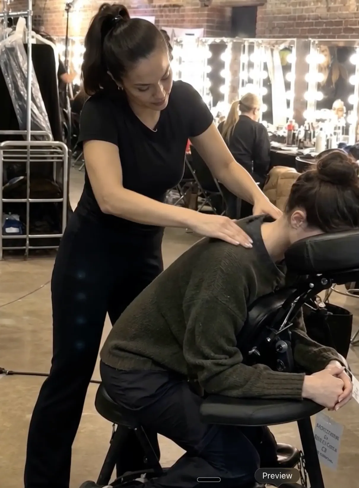 Knead NYC therapist providing backstage chair massage beside illuminated dressing-room mirrors
